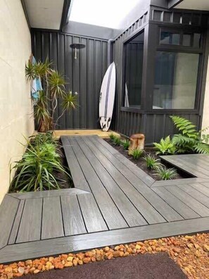 Una moderna veranda esterna con un pavimento in legno, piante in vaso e una tavola da surf appoggiata al muro.