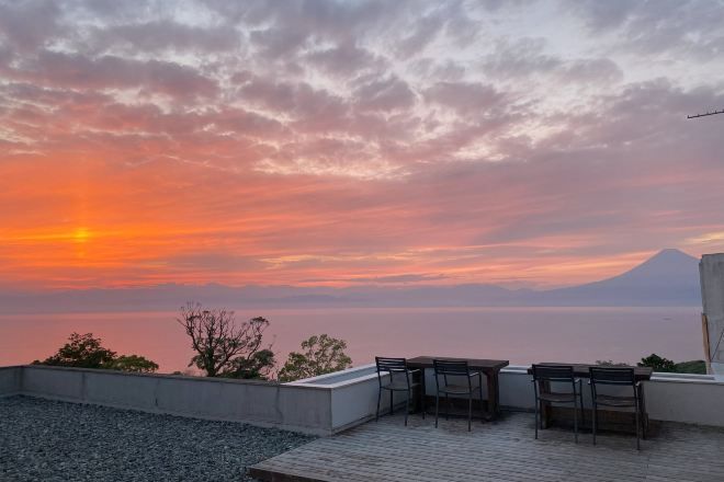 Vista do pôr do sol a partir de um terraço com mesa e cadeiras, com vista para um corpo d'água e uma montanha.