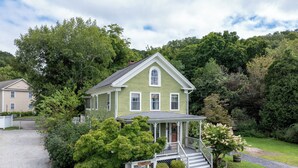 Une maison à deux étages avec un porche, entourée d’une végétation luxuriante et d’arbres.