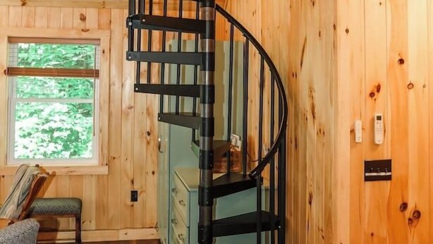 Ein Holzinnenraum mit einer Wendeltreppe, einem Fenster mit Blick auf Grünflächen und einem Bett mit Kopfteil.