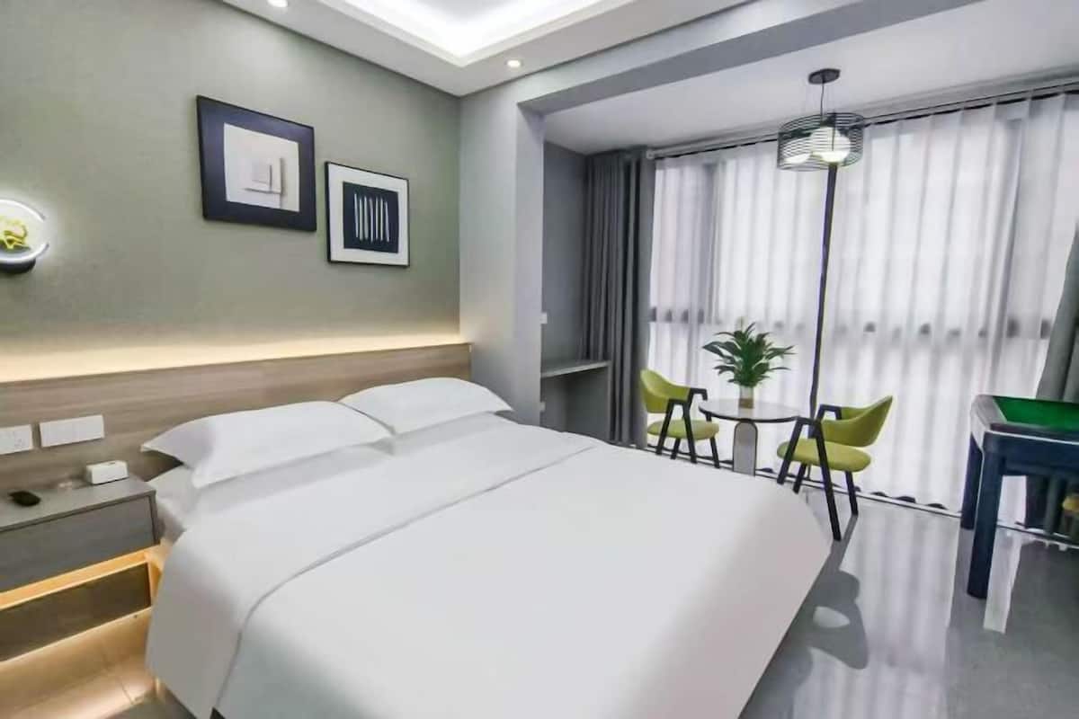 A modern hotel room with a large bed, bedside tables, a desk with chairs, and a view of the outside through large windows.