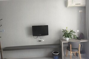 A modern room with a flat-screen TV mounted on the wall, a small desk with a chair, a potted plant, and a trash bin. - Tianshun Apartment (Chongqing)