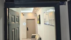 Ένας διάδρομος με μια κλειστή πόρτα, μια μικρή περιοχή με καθίσματα και ένα μαξιλάρι, και ένας πίνακας πληροφοριών τοποθετημένος στον τοίχο.