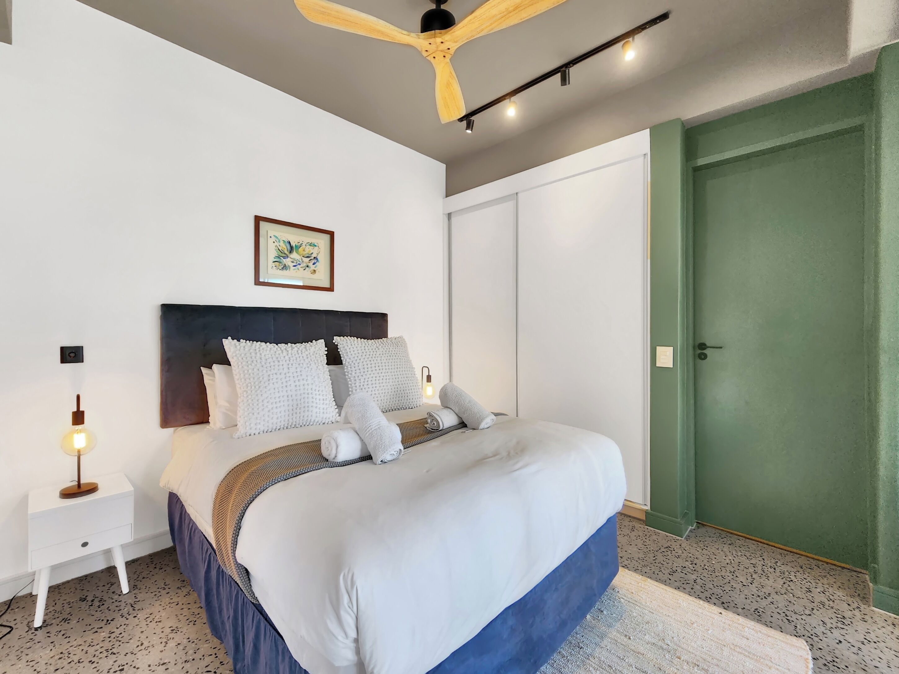 A bedroom with a bed, a nightstand, a ceiling fan, a wall-mounted light, and a green door.
