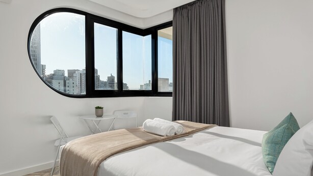 Habitación de hotel moderna con un ventanal grande, una cama con sábanas blancas, una mesita con una planta y una silla.