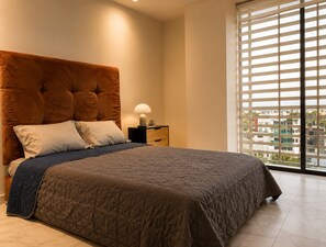 A bedroom with a large bed, a nightstand, a lamp, and a view of buildings through the window.