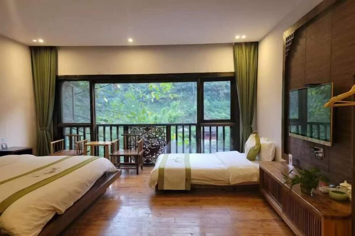 A hotel room with two beds, a TV, a desk, and a balcony with a view of greenery.
