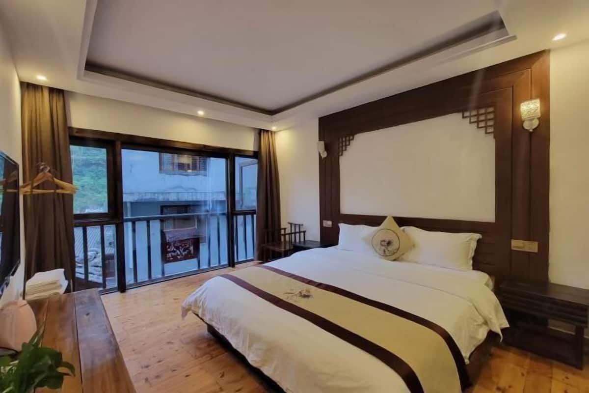 A hotel room with a large bed, wooden flooring, a balcony with a view, and a wooden headboard.