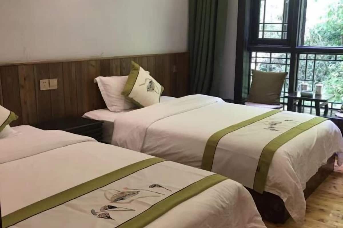 A hotel room with two beds, a wooden headboard, a window with a view of greenery, and a small table with a chair.