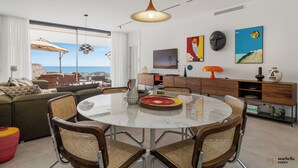 A modern living room with a round glass table, wicker chairs, a sofa, and a view of the sea.
