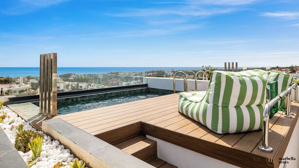 A rooftop pool with a wooden deck, green-striped cushions, and a clear view of the sea and cityscape.