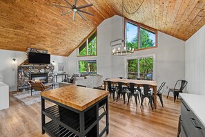 Un loft espacioso con techo alto, una chimenea de piedra, una zona de comedor con una larga mesa de madera y grandes ventanales que ofrecen vistas al exterior.