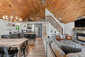 A spacious living area with a high ceiling, wooden beams, a central staircase, a kitchen with modern appliances, and a fireplace.