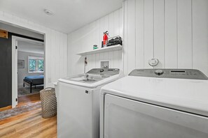 A laundry room with two front-loading washers and dryers, a shelf with cleaning supplies, and a door leading to another room.