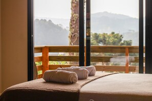 A bed with a beige bedspread and two rolled white towels on it, positioned in front of a window with a view of a wooden railing and greenery.
