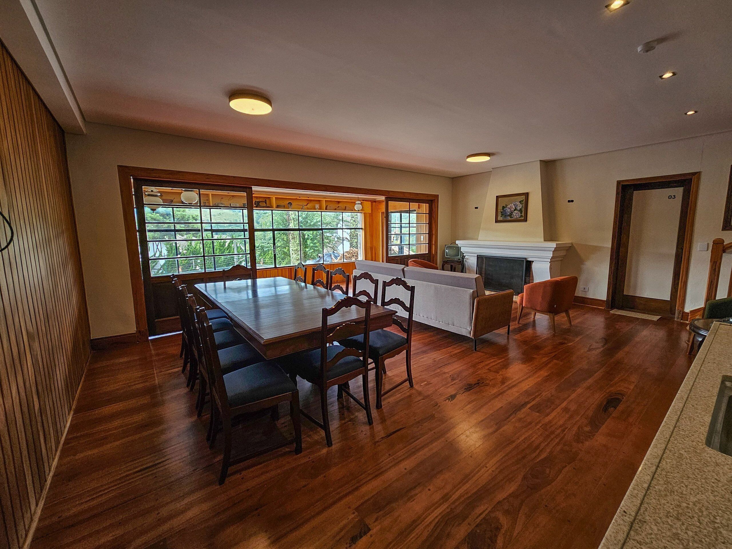 Um quarto espaçoso com uma grande mesa de jantar de madeira, várias cadeiras, uma lareira e vista através de amplas janelas.