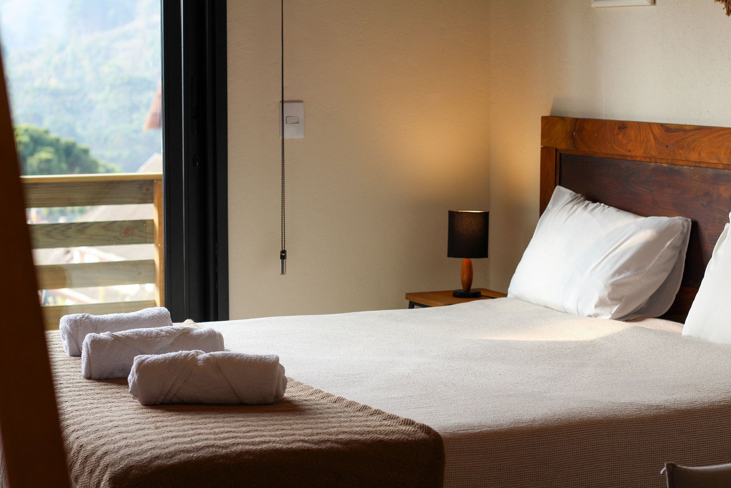 A neatly made bed with white linens and a wooden headboard, a bedside lamp, and a view of a balcony with a wooden railing.