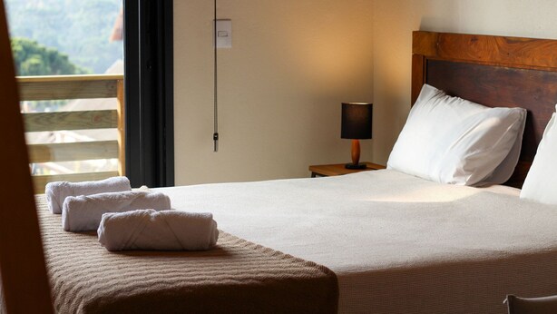 A neatly made bed with white linens and a wooden headboard, a bedside lamp, and a view of a balcony with a wooden railing. - Estudios no Capivari by The Lodges (Campos do Jordão)