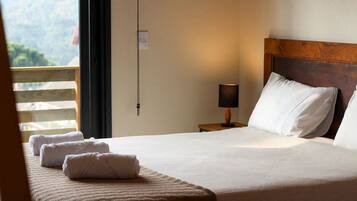 A neatly made bed with white linens and a wooden headboard, a bedside lamp, and a view of a balcony with a wooden railing.