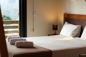 Een net opgemaakt bed met witte lakens en een houten hoofdbord, een bedlampje, en uitzicht op een balkon met houten leuning.