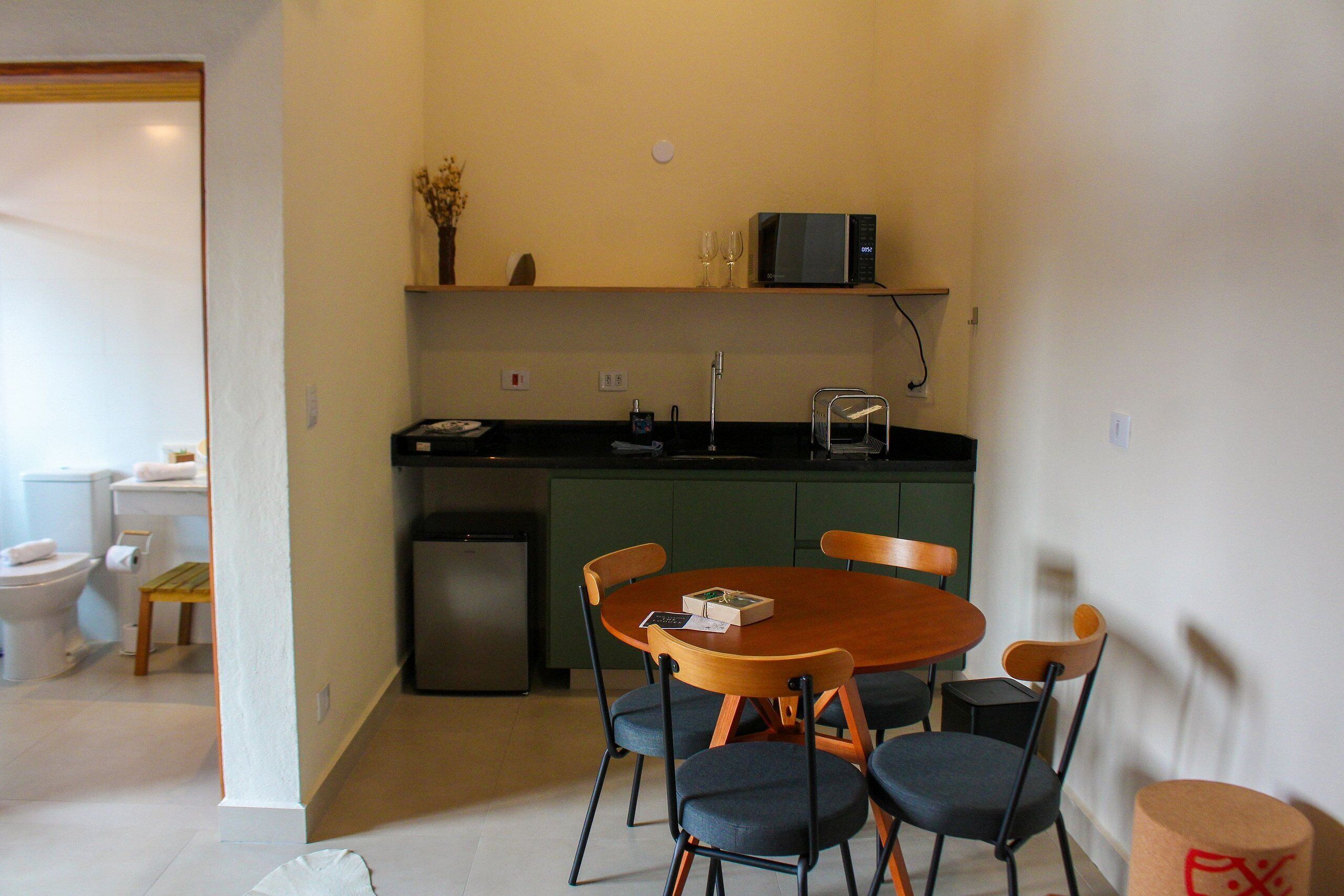 A compact kitchen with a microwave, sink, and a small refrigerator. There is a dining table with four chairs and a vase with dried flowers on the counter.