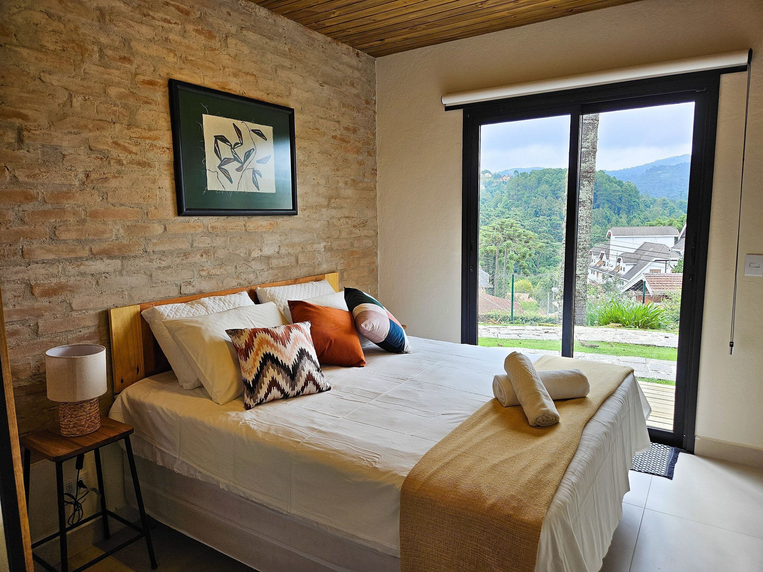 A bedroom with a large bed, a wooden headboard, a brick wall, a framed picture, and a view of a green landscape through a large window.