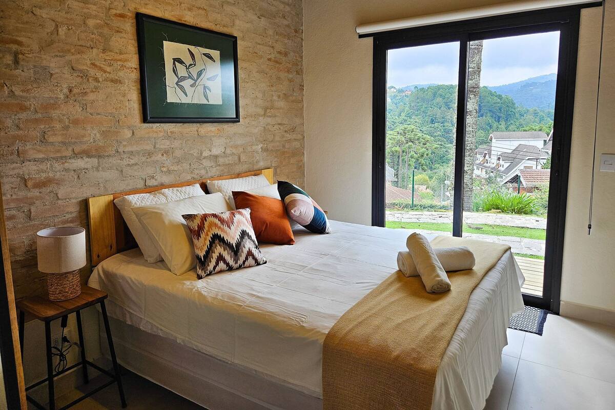A bedroom with a large bed, a wooden headboard, a brick wall, a framed picture, and a view of a green landscape through a large window.