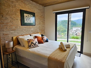 Een slaapkamer met een groot bed, een houten hoofdeinde, een bakstenen muur, een lijst met een afbeelding en uitzicht op een groen landschap door een groot raam.