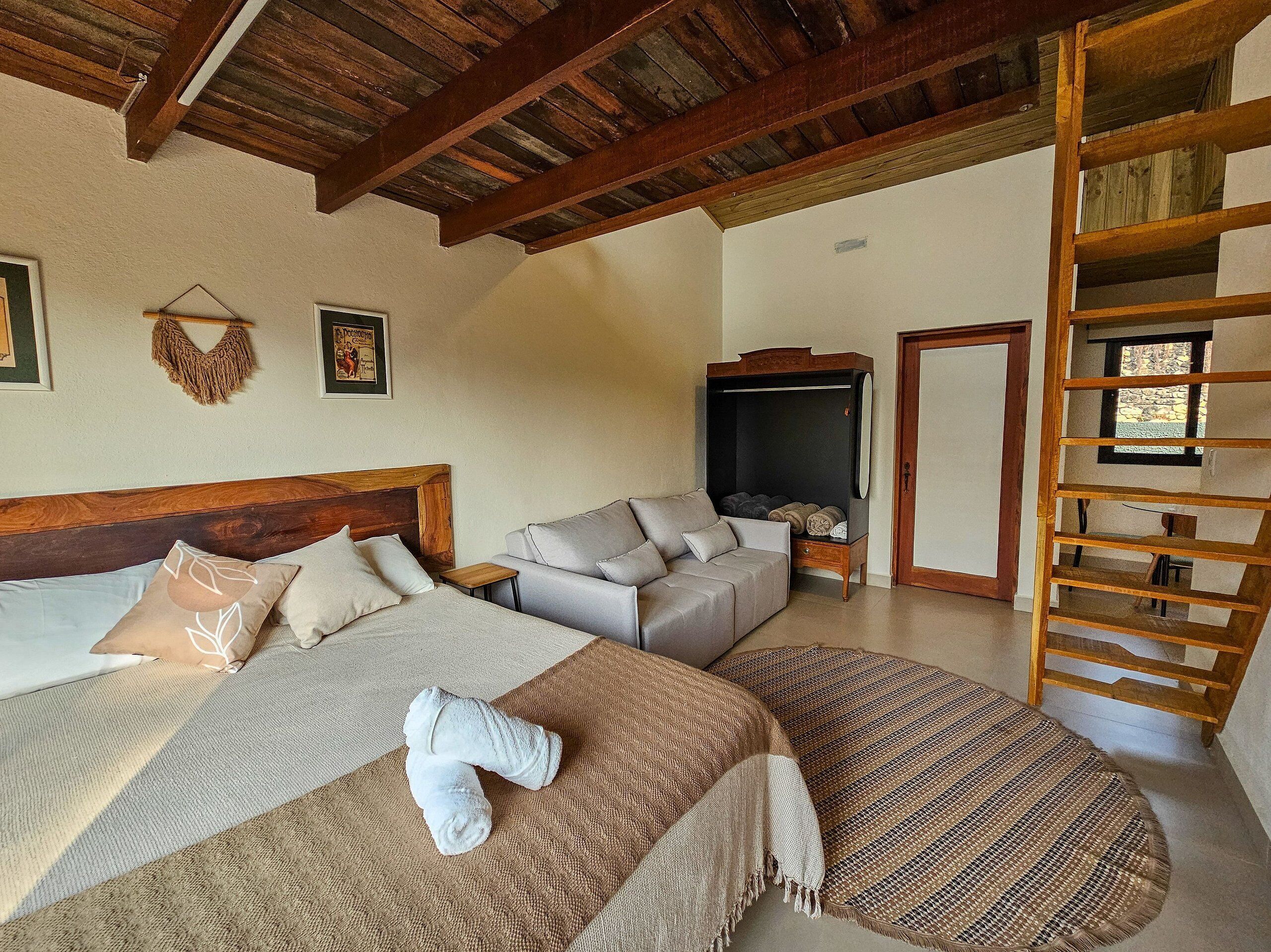 Une chambre avec un plafond en bois, un lit avec une tête de lit, un canapé, un meuble pour la télévision et une échelle en bois.
