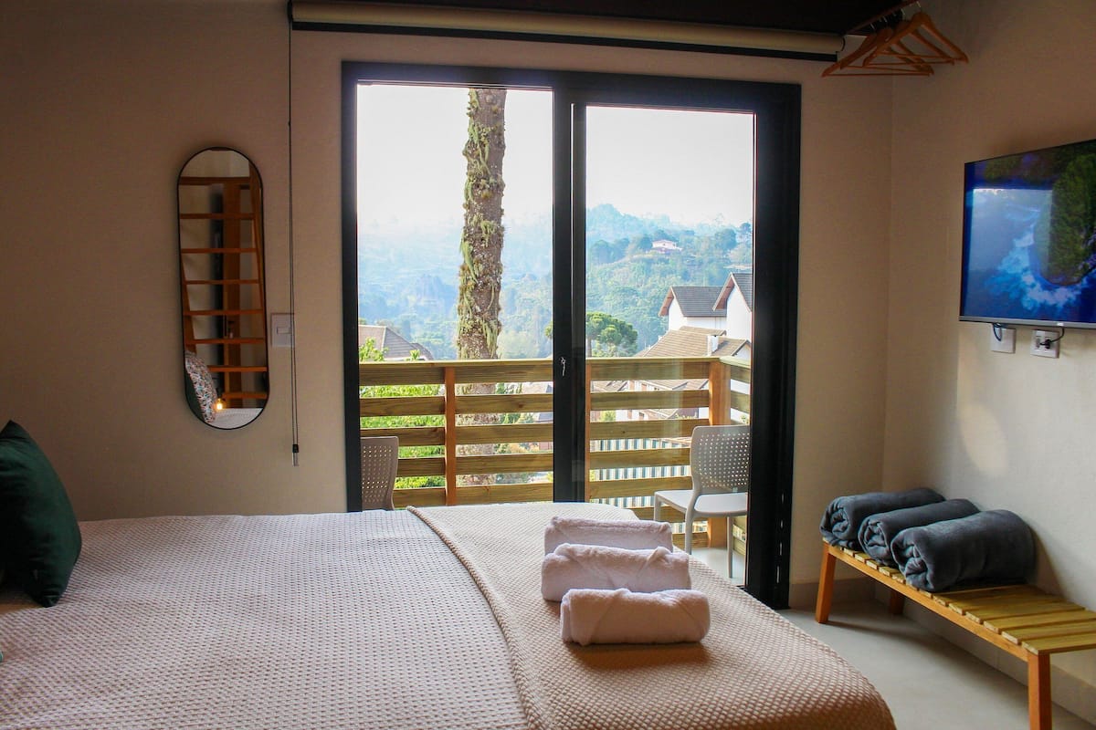 A bedroom with a large bed, a wooden bench with towels, a TV, and a balcony with a view of the mountains.