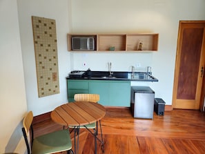 Een kleine keuken met een magnetron, spoelbak en kleine koelkast. Er is een houten tafel met twee stoelen en een decoratief wandobject.