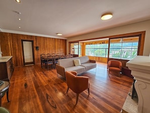 Een ruime kamer met houten vloeren, een bank, twee fauteuils, een eettafel met stoelen, en grote ramen met uitzicht op groen.