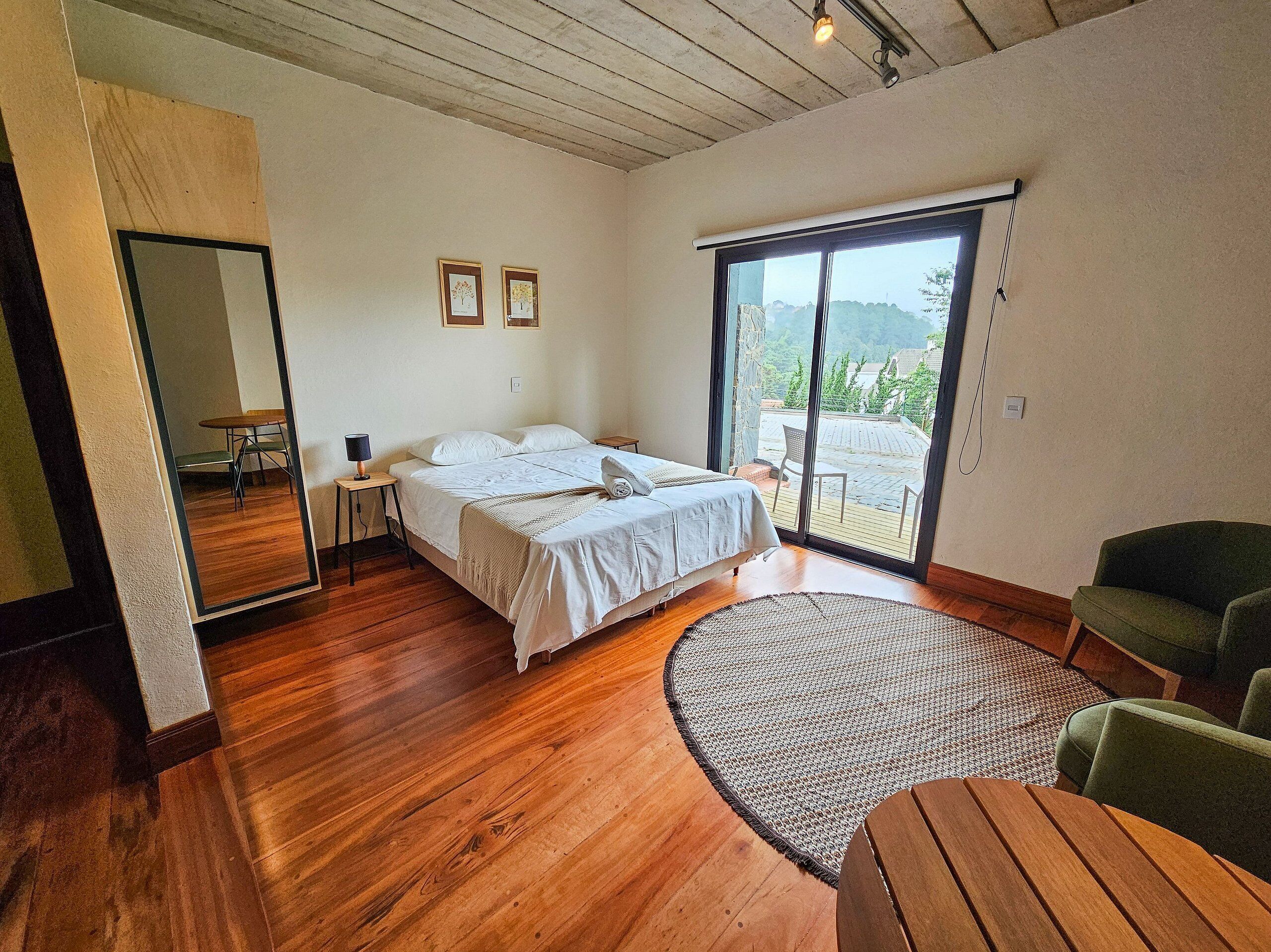 Quarto com cama, poltrona verde, mesa redonda de madeira e uma janela grande com vista para o exterior.