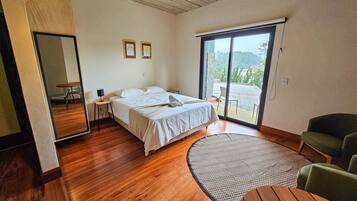 Quarto com cama, poltrona verde, mesa redonda de madeira e uma janela grande com vista para o exterior.