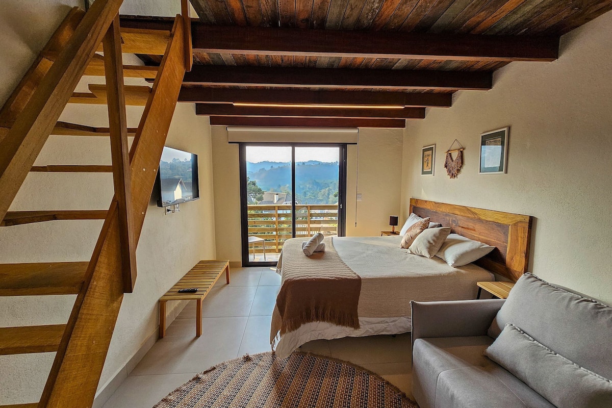 A bedroom with a wooden staircase, a bed with pillows, a sofa, and a television on the wall.