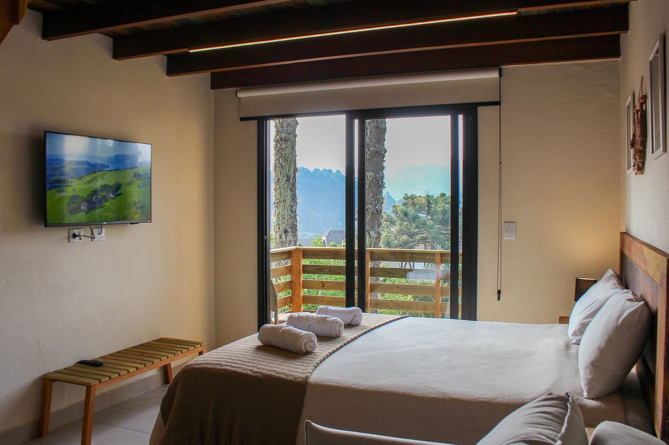 A bedroom with a large bed, a bench, a TV mounted on the wall, and a balcony with a view of the mountains.