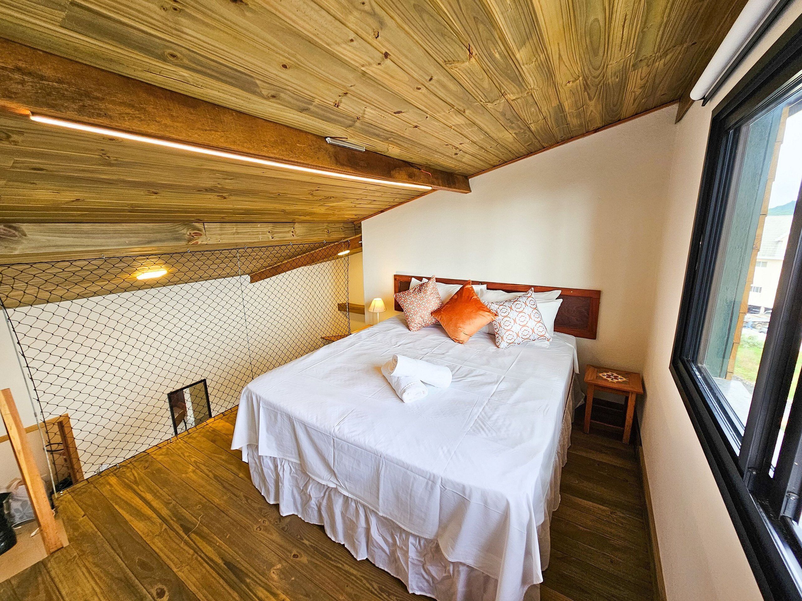 A bedroom with a wooden ceiling, a bed with white bedding, and a view through a window.