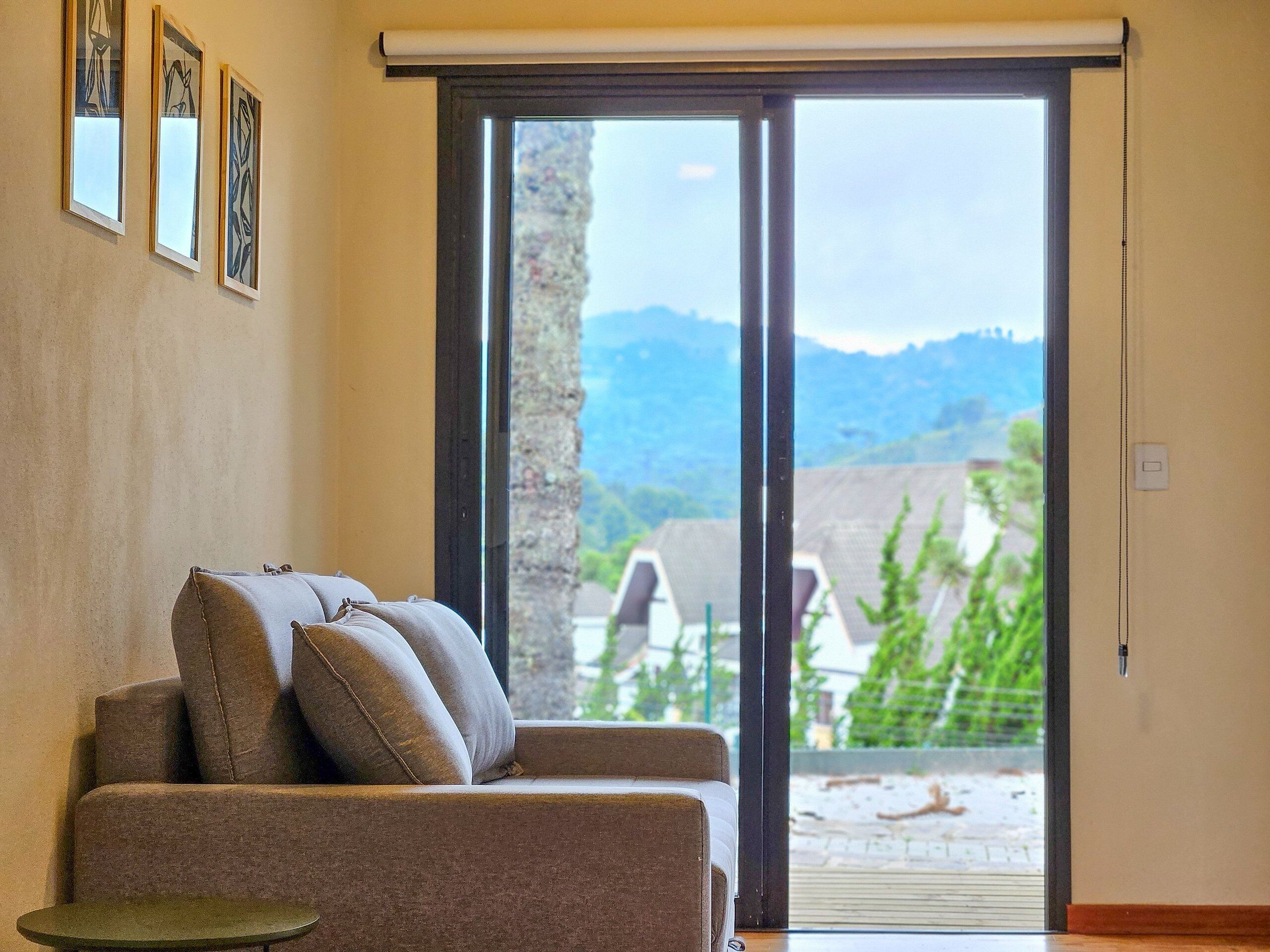 Sala de estar com sofá, mesa pequena e porta de vidro deslizante que dá para área externa com vista para montanhas e vegetação.