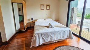 A bedroom with a large bed, a wooden floor, a mirror, and a view of the outdoors.