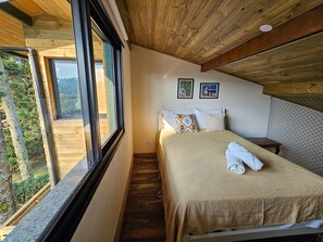 Een slaapkamer met een groot raam, houten plafond en een bed met een beige deken en twee kussens.