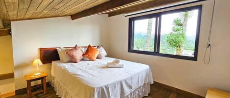 Un dormitorio con techo de madera, un ventanal con vistas, una cama con ropa blanca y una mesita de noche con una lámpara.