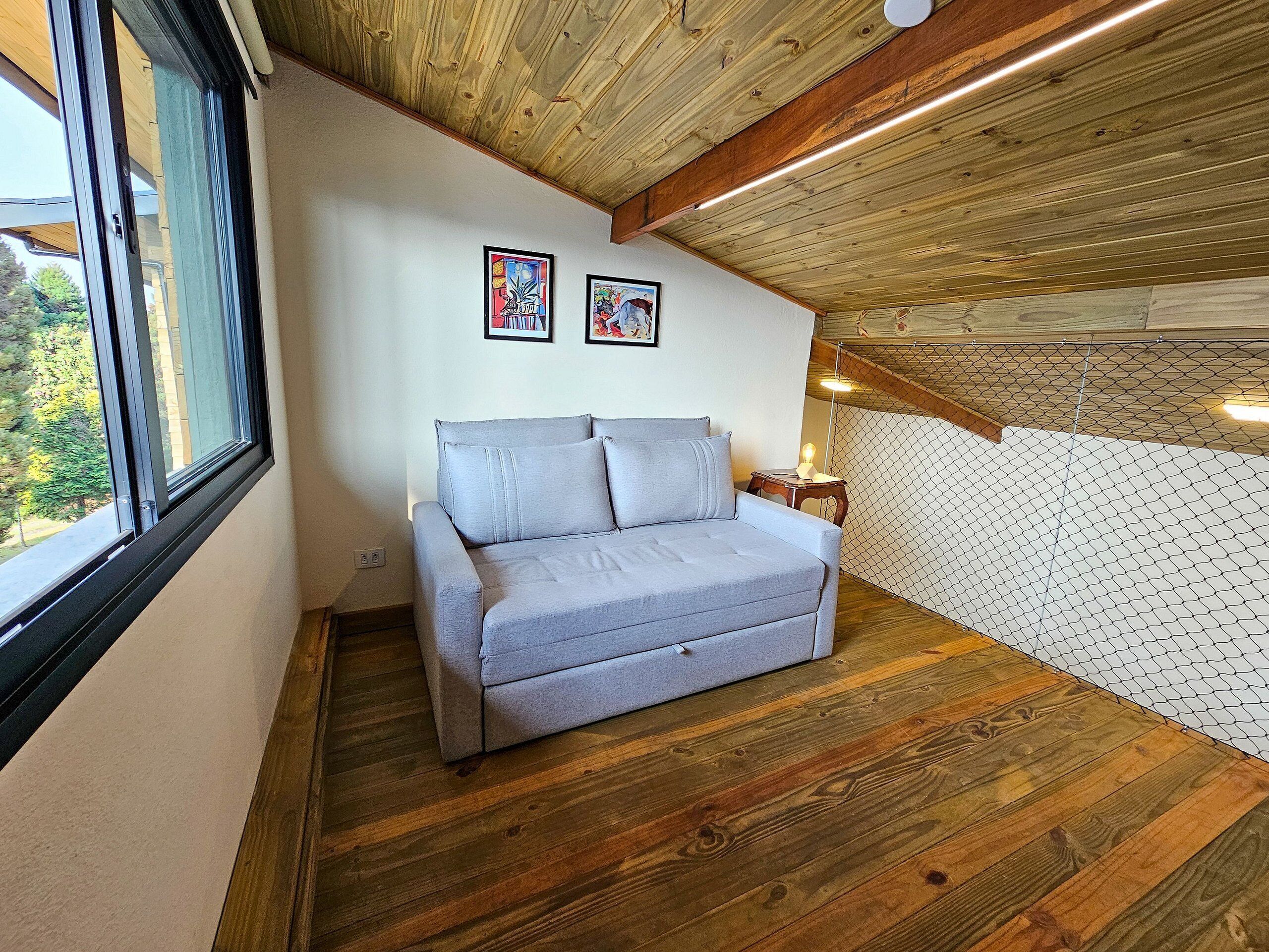 Una habitación acogedora con un sofá gris, suelo de madera, techo con vigas a la vista y una ventana.