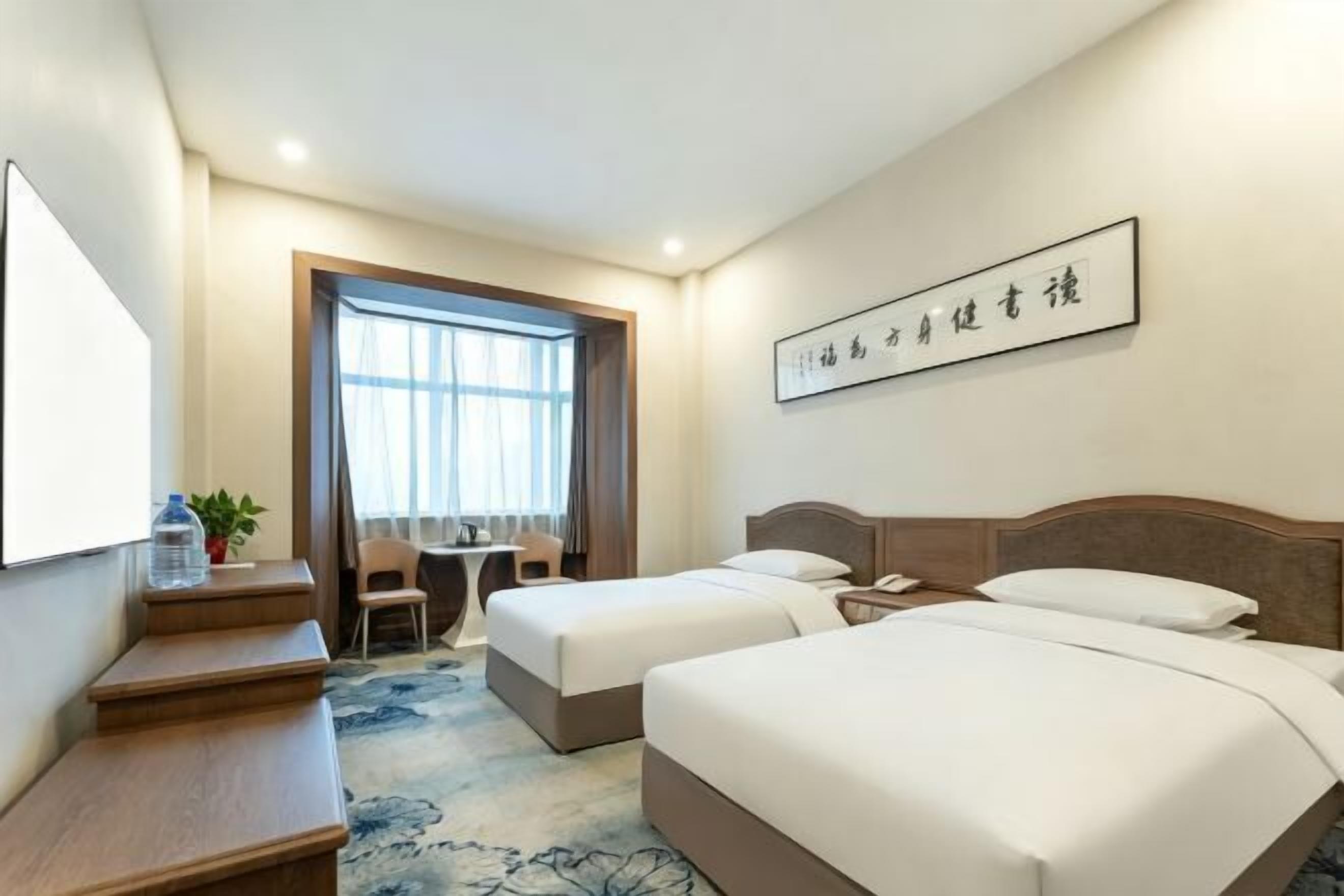 A hotel room with two beds, a desk, a chair, a small table, and a window with curtains.