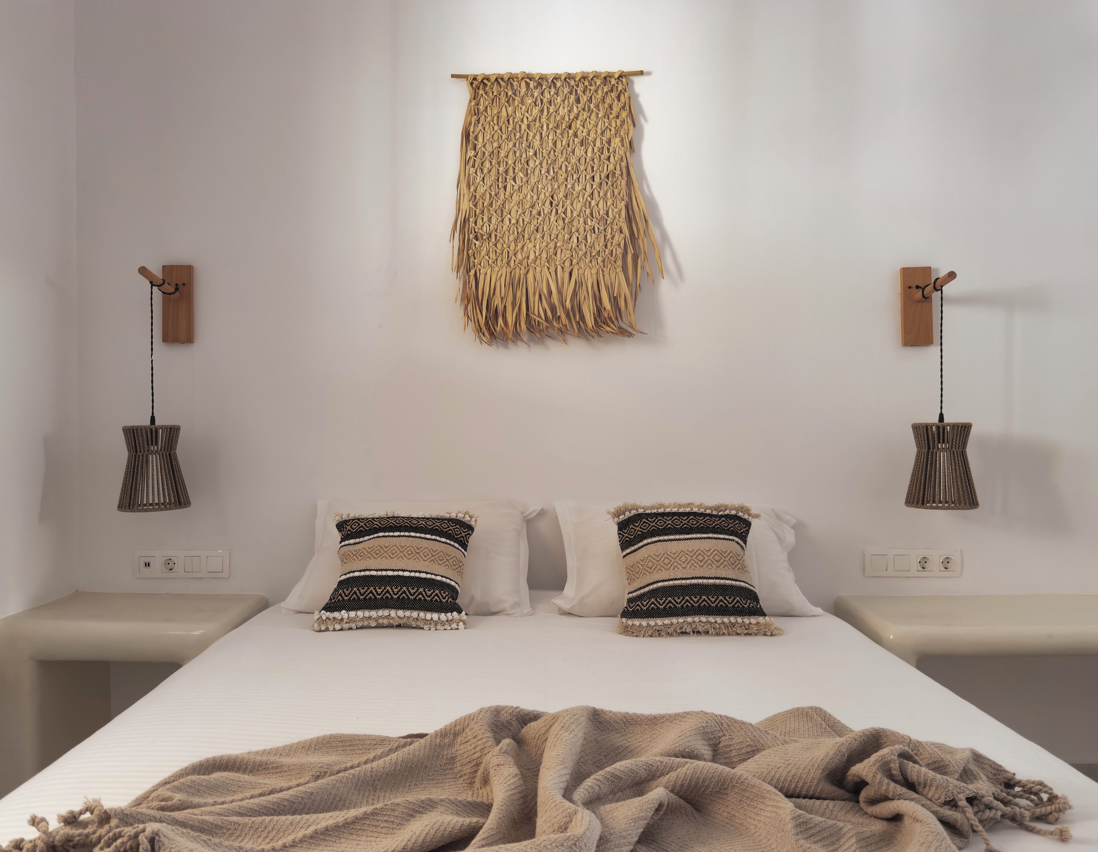 Ein ordentlich eingerichtetes Schlafzimmer mit einem Bett, zwei Nachttischen, einem gewebten Wandbehang und zwei Pendelleuchten.