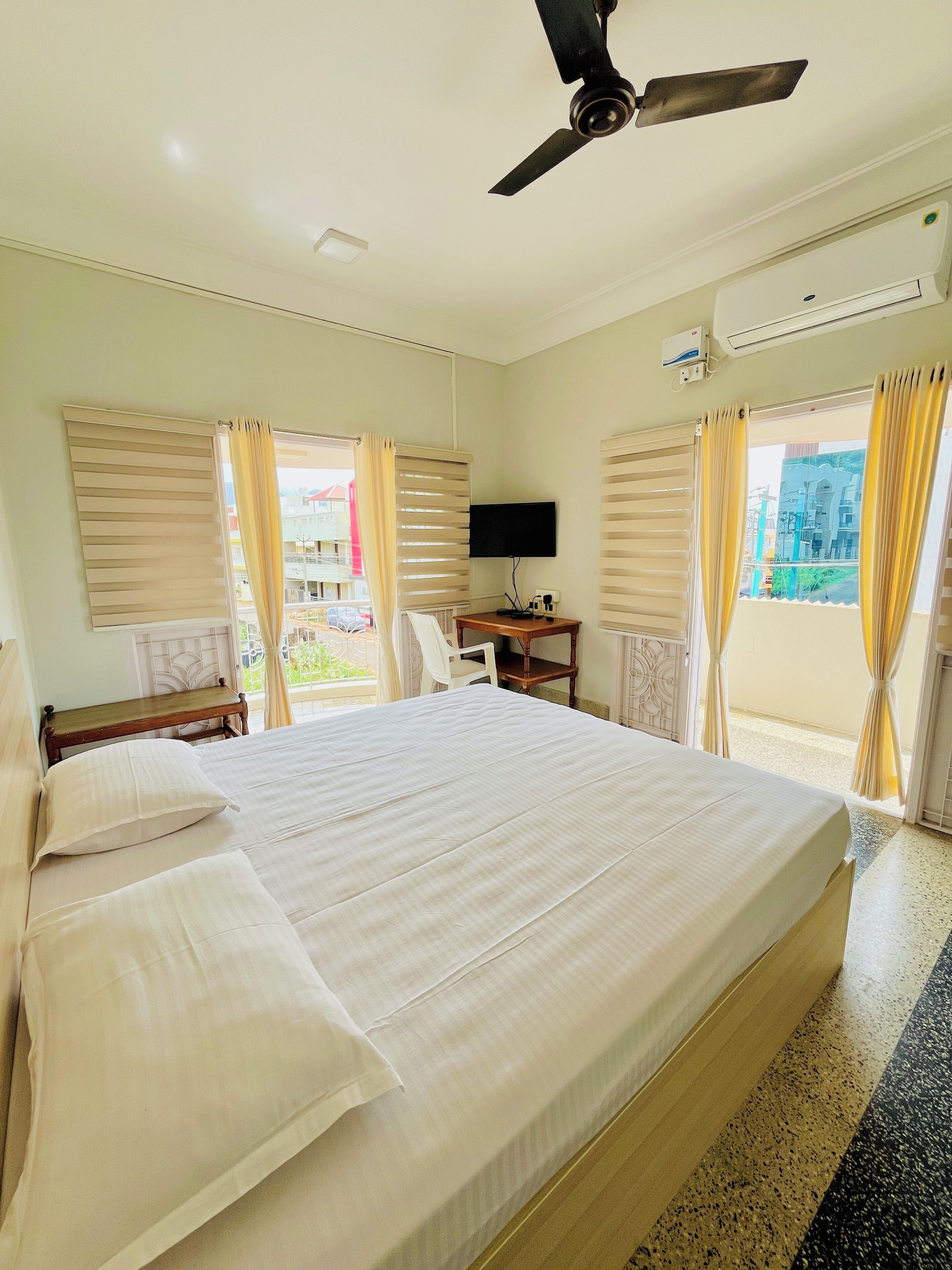 Une chambre d’hôtel avec un grand lit, un bureau avec un ordinateur, un ventilateur de plafond et des fenêtres équipées de rideaux.