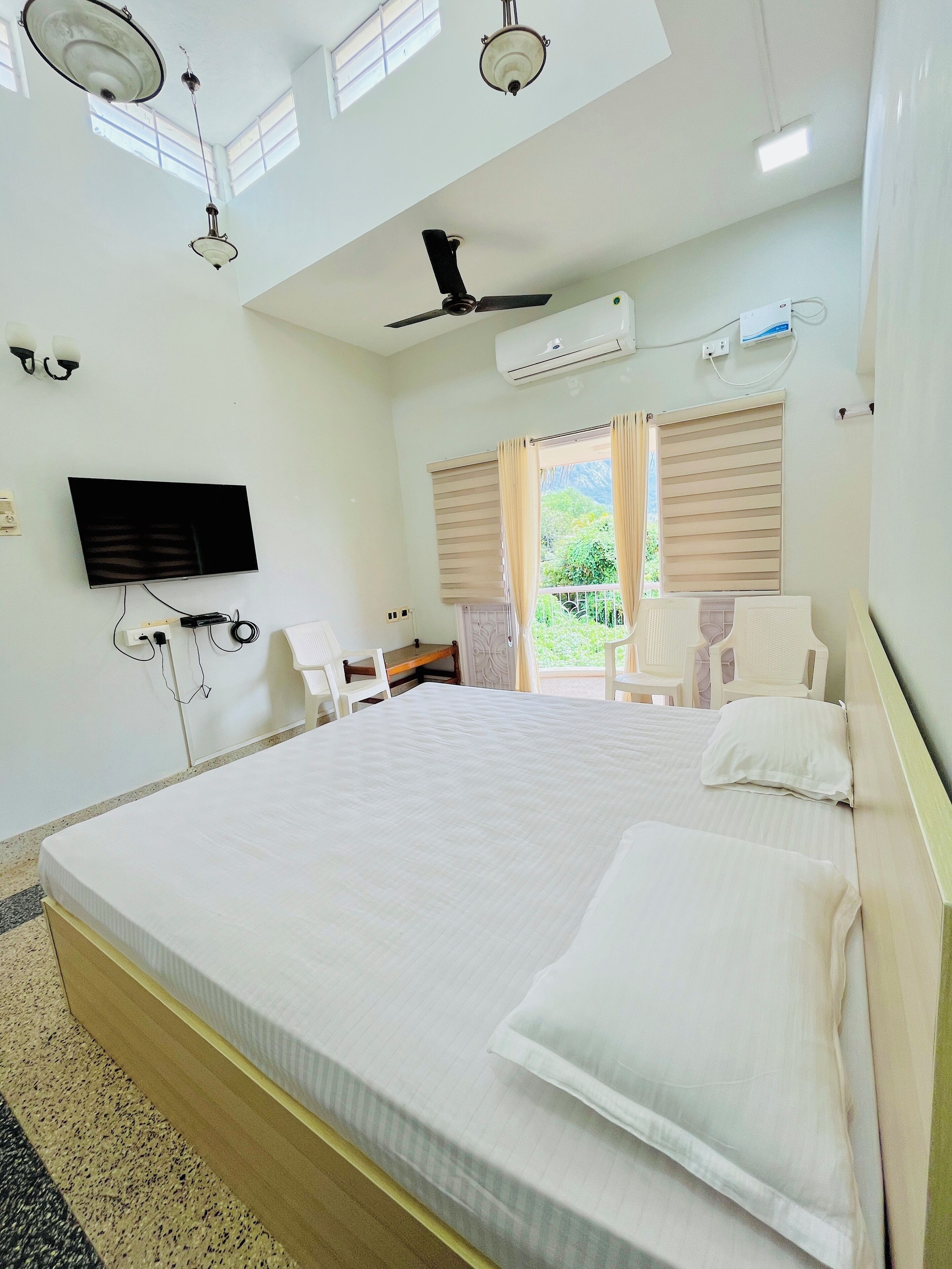 A hotel room with a large bed, ceiling fans, an air conditioning unit, a flat-screen TV, and a view of greenery outside.