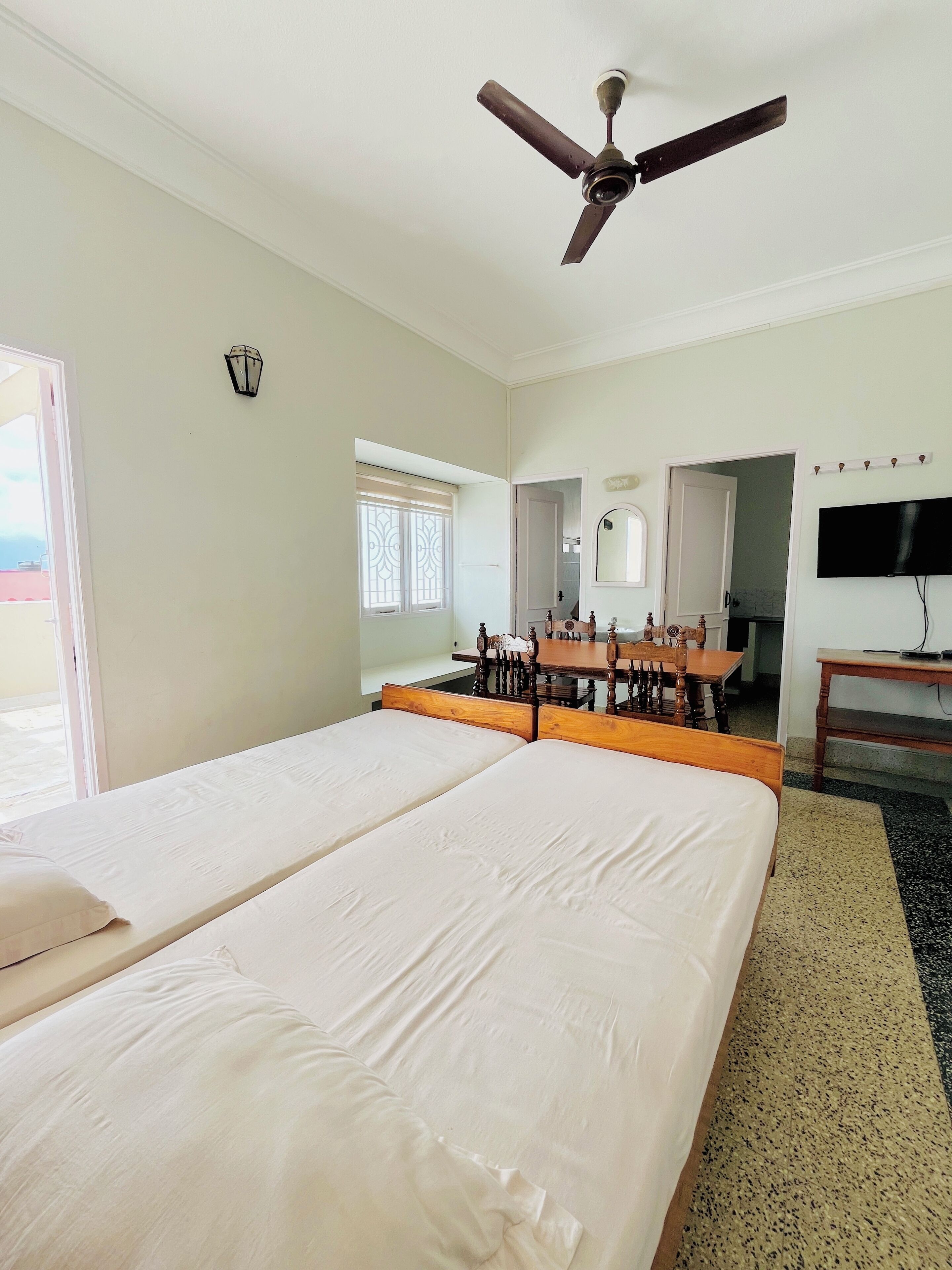 A hotel room with a large bed, a ceiling fan, a dining table with chairs, a TV, and a window with blinds.