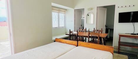 A hotel room with a large bed, a ceiling fan, a dining table with chairs, a TV, and a window with blinds.