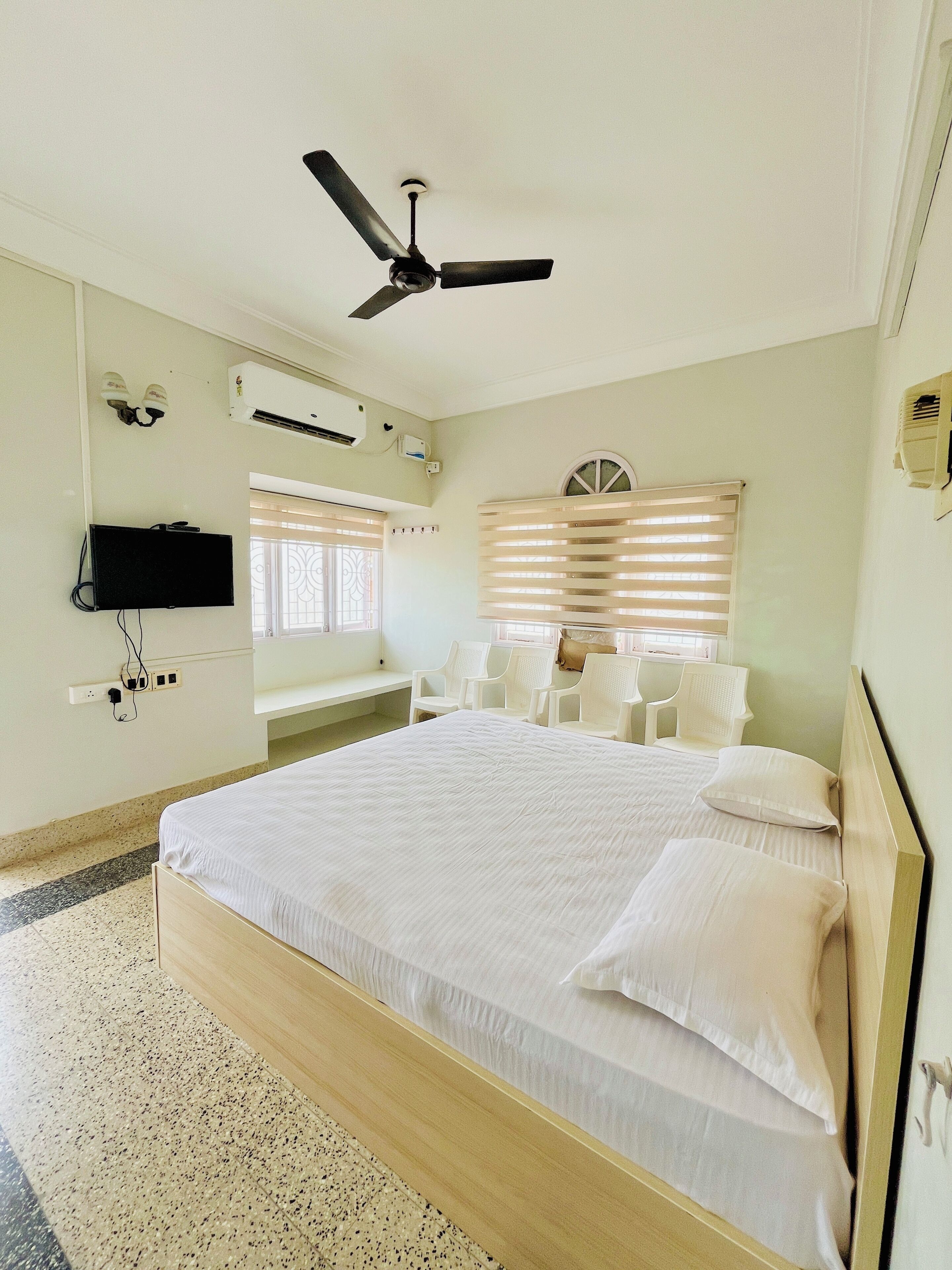 A bedroom with a bed, ceiling fan, air conditioning unit, TV, and a window with blinds.