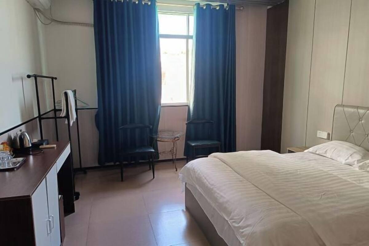 Ein Hotelzimmer mit einem Bett, einem Schreibtisch mit Wasserkocher, zwei Stühlen, einem kleinen Tisch und einem Fenster mit Vorhängen.
